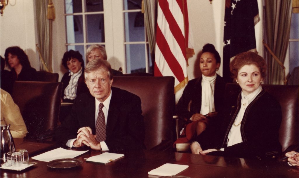 Dr. Sarah Weddington seated to the left of Pres. Carter, with other staff members seated behind them and on either side.